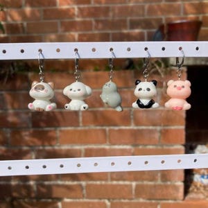 May include: Five small animal charms hanging from a white display bar. The charms include a brown and white cat, a white dog, a grey seal, a black and white panda, and a pink pig. Each charm is attached to a silver-toned metal clasp.