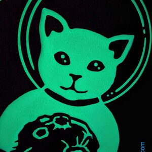 K&ouml;nnte beinhalten: Eine gr&uuml;n leuchtende Katze, die einen Astronautenhelm tr&auml;gt und einen Mondstein h&auml;lt. Die Katze l&auml;chelt und hat gro&szlig;e Augen.