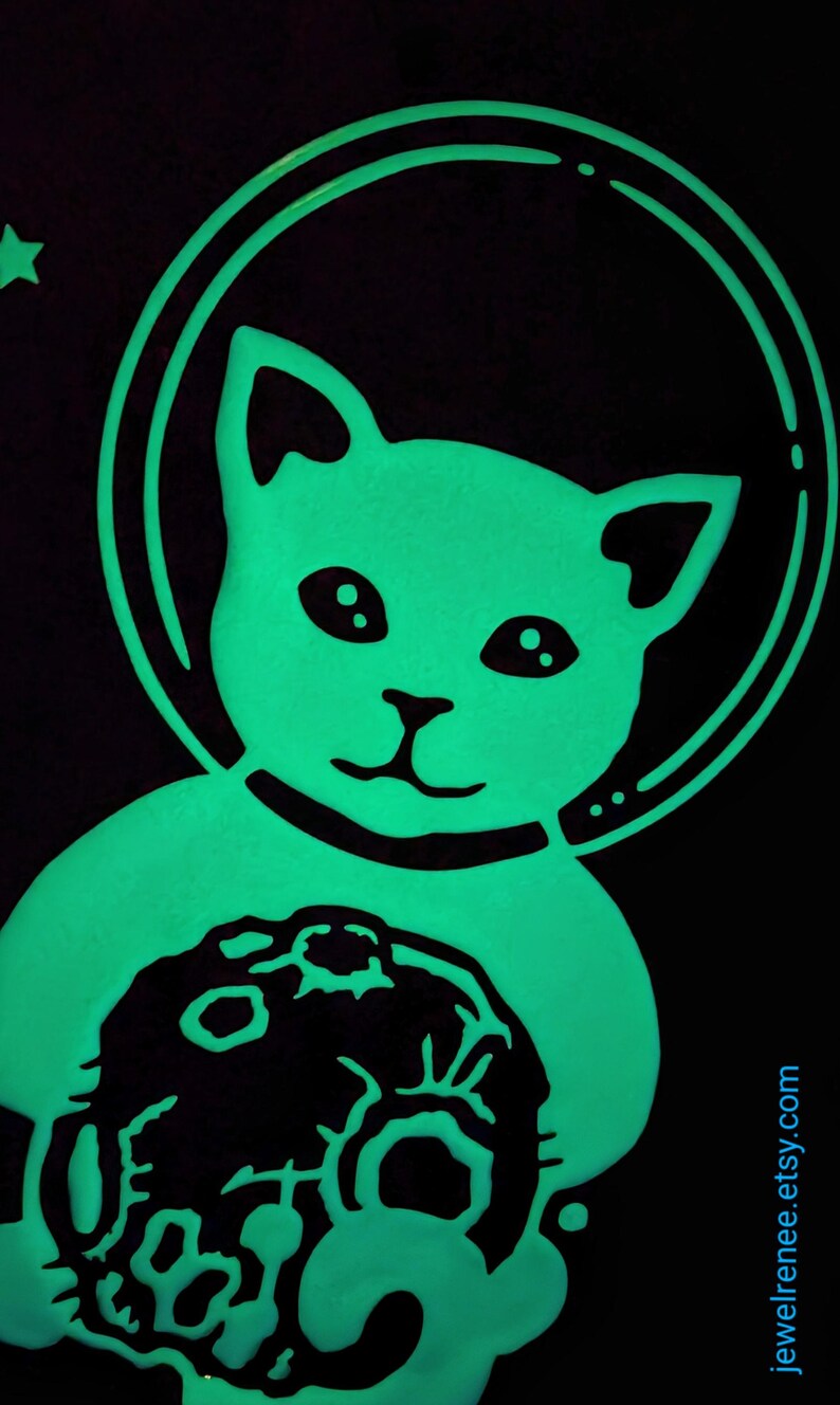 K&ouml;nnte beinhalten: Eine gr&uuml;n leuchtende Katze, die einen Astronautenhelm tr&auml;gt und einen Mondstein h&auml;lt. Die Katze l&auml;chelt und hat gro&szlig;e Augen.