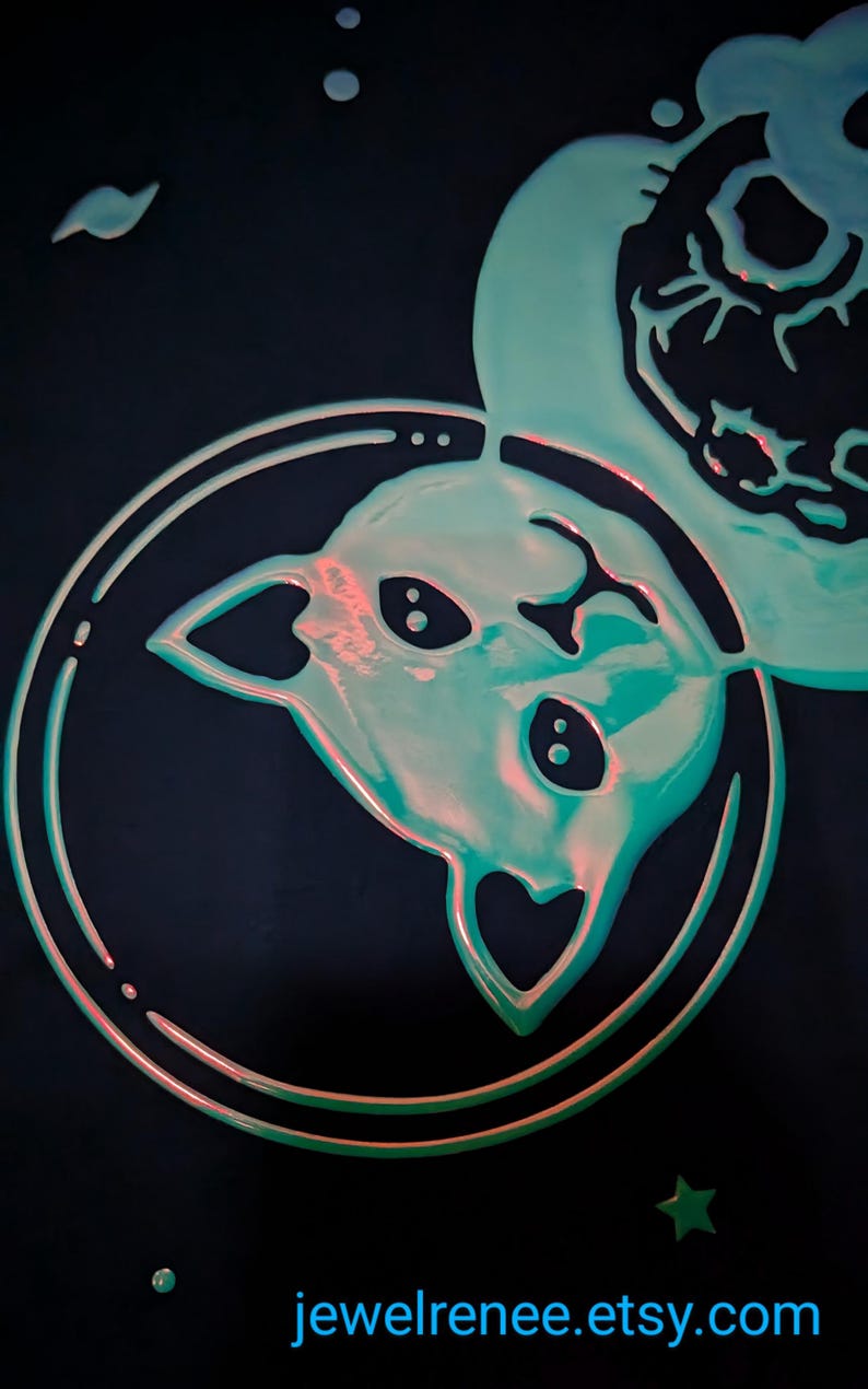 K&ouml;nnte beinhalten: Eine gr&uuml;ne und rosafarbene Katze-Astronauten-Illustration mit einem herzf&ouml;rmigen Kopf, die im Weltraum mit einem Stern und einem Planeten im Hintergrund schwebt. Die Illustration befindet sich auf einem schwarzen Hintergrund. Der Text "jewelrenee.etsy.com" befindet sich am unteren Rand des Bildes.