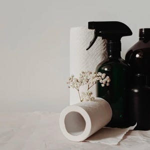 Puede incluir: Un bodegón con productos de limpieza. Un pulverizador verde oscuro, una botella negra y una botella negra pequeña se agrupan con un rollo de toallas de papel y un rollo de papel higiénico. Flores blancas se colocan en el rollo de papel higiénico.