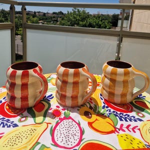 Peut inclure: Trois mugs en céramique avec un motif à rayures orange et blanc sont présentés sur une nappe colorée. Les mugs ont des intérieurs bruns et des anses incurvées. L'arrière-plan montre un ciel bleu clair et une balustrade de balcon.