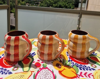 May include: Three ceramic mugs with a striped design in shades of orange and white are displayed on a colorful tablecloth. The mugs have brown interiors and curved handles. The background shows a clear blue sky and a balcony railing.
