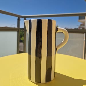 Könnte beinhalten: Hoher Keramik-Krug mit schwarz-weißen Streifen. Der Krug hat einen gebogenen Griff und einen Ausguss. Er steht auf einer gelben Oberfläche, mit einem hellblauen Himmel im Hintergrund.