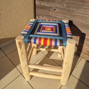 May include: A small wooden stool with a woven seat. The seat has a colourful geometric pattern with blue, yellow, red, and black threads. The legs are made of light-coloured wood.