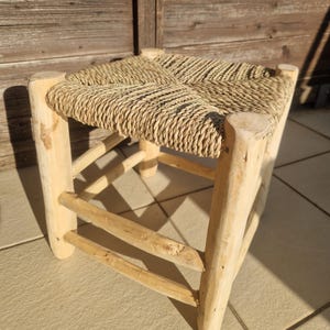 Puede incluir: Un pequeño taburete cuadrado con un asiento tejido de color marrón claro y patas de madera clara. Las patas están conectadas por soportes horizontales de madera. El taburete está hecho de materiales naturales y tiene un aspecto rústico.