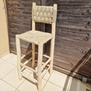 Op de afbeelding: Een hoge houten barkruk met een geweven zitting en rugleuning. De kruk heeft vier poten en een voetensteun. De rugleuning is rechthoekig en de zitting is vierkant. Het hout heeft een natuurlijke, onafgewerkte uitstraling. Ongeveer 100 cm hoog.