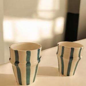 Peut inclure: Deux tasses en céramique blanche avec des rayures verticales bleues. Les tasses sont légèrement évasées en haut et en bas.