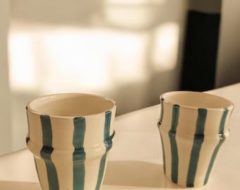 May include: Two white ceramic cups with blue vertical stripes. The cups are slightly flared at the top and bottom.