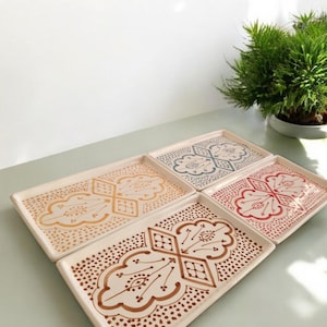 Peut inclure: Ensemble de six plateaux rectangulaires en céramique avec des motifs décoratifs. Chaque plateau présente une base crème avec un motif de couleur différent, notamment marron, jaune, bleu et rouge. Les plateaux sont disposés sur une surface vert clair.