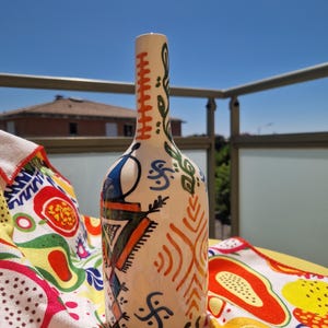Peut inclure: Une bouteille en céramique blanche ornée de motifs abstraits colorés en bleu, orange, vert et noir. La bouteille a un long col et est décorée de formes géométriques et organiques. La bouteille est posée sur un tissu à motifs colorés.