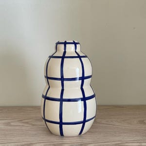 Peut inclure: Un vase en céramique crème avec un motif quadrillé bleu. Le vase a une forme arrondie avec un col étroit et une base plus large. Les lignes bleues créent un design géométrique, ajoutant une touche moderne à la pièce décorative.