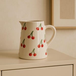 Op de afbeelding: Een crèmekleurige keramische kan versierd met een herhalend patroon van rode kersen met groene stelen. De kan heeft een gebogen handvat en een tuit om uit te schenken. Het ontwerp is eenvoudig en charmant, geschikt voor het serveren van dranken.
