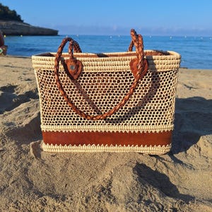 Könnte beinhalten: Gewebte Stroh-Tragetasche mit rechteckiger Form, mit braunem Lederboden und geflochtenen Griffen. Die Tasche hat ein helles, offenes Webdesign, perfekt für den Transport von Strandutensilien.