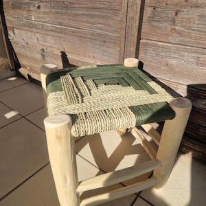 Può includere: Un piccolo sgabello in legno fatto a mano con un sedile intrecciato. Il sedile presenta un motivo di materiale intrecciato verde e color naturale. Le gambe sono in legno chiaro e lo sgabello ha un design semplice e rustico. Lo sgabello è alto circa 30 cm.