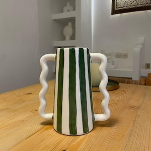 Peut inclure: Un vase en céramique à rayures blanches et vertes avec deux poignées ondulées. Le vase est cylindrique avec un dessus évasé et repose sur une table en bois. Les rayures vertes sont verticales et espacées uniformément.