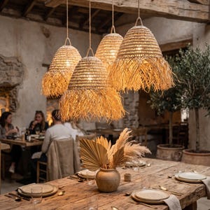 Peut inclure: Un ensemble de quatre suspensions en paille tressée avec des bords effilochés suspendues à une poutre en bois. Les luminaires présentent une forme conique de couleur beige naturel et sont suspendus par de fines cordes au-dessus d'une table.