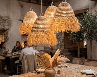 Puede incluir: Un conjunto de cuatro lámparas colgantes de paja tejida con bordes deshilachados que cuelgan de una viga de madera. Las luminarias tienen una forma cónica de color beige natural y están suspendidas por cuerdas finas sobre una mesa larga.