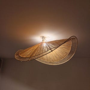 Peut inclure: Un plafonnier tissé de couleur naturelle. Le luminaire a un abat-jour large et incurvé en paille tressée ou matériau similaire, avec un bord décoratif. Il est monté sur une base blanche.