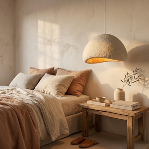 Könnte beinhalten: Ein Schlafzimmer mit einem Bett mit Bettwäsche in neutralen Tönen, darunter Kissen und eine Decke in Beige- und Rosttönen. Ein rustikaler Nachttisch aus Holz beherbergt Bücher, ein Tablett und eine Vase mit Grünzeug. Eine große, strukturierte Pendelleuchte hängt darüber.