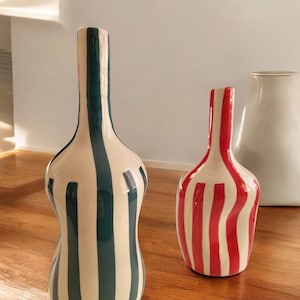 May include: Two decorative ceramic vases with vertical stripe patterns. One vase is tall with dark teal stripes, the other is shorter with red stripes. Both vases have a cream-colored background and are on a wooden surface.