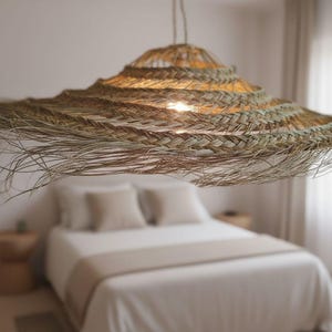 Puede incluir: Una lámpara colgante grande, tejida con fibras naturales, cuelga sobre una cama. La luz tiene un diseño amplio y escalonado con un brillo cálido. El dormitorio presenta una paleta de colores neutros.
