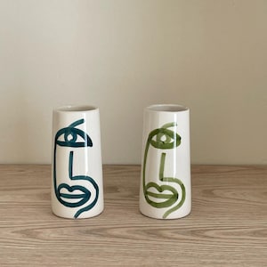 Peut inclure: Deux vases en céramique blanche avec des motifs de visages abstraits. L'un présente un contour de visage bleu-vert, l'autre un visage vert. Les vases cylindriques sont posés sur une surface en bois.