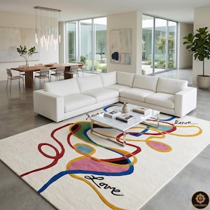 Könnte beinhalten: Ein modernes Wohnzimmer mit einem großen weißen Ecksofa, einem Couchtisch mit Marmorplatte und einem farbenfrohen abstrakten Teppich. Der Teppich hat einen cremefarbenen Hintergrund mit roten, blauen, gelben und rosa Wirbeln und dem Wort "Love" in schwarzer Schrift.
