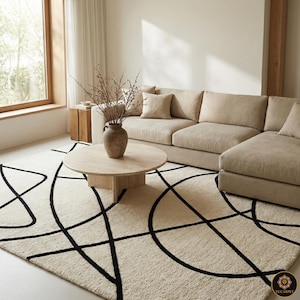 May include: A beige sectional sofa and a round wooden coffee table are in a well-lit living room. A cream-colored rug with a black geometric design covers the floor. A vase with dried branches sits on the table. A wooden side table is near a large window.