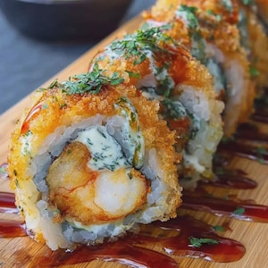 May include: Close-up of a sushi roll with a fried exterior, filled with shrimp and a creamy, green-tinted filling. The roll is drizzled with a dark sauce and garnished with fresh herbs. The sushi is presented on a wooden board.