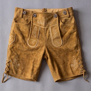 May include: Tan suede leather shorts featuring a drop-front flap, light-colored decorative embroidery on the front panels and legs, metal buttons at the waistband, and side lacing details with matching leather cords at the bottom of each leg.