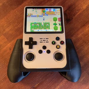 Può includere: Una console di gioco portatile bianca con pulsanti neri e uno schermo che mostra un gioco di simulazione di fattoria. La console ha due controller neri attaccati ai lati.