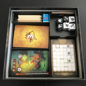 Könnte beinhalten: Ein Brettspiel-Organizer mit Fächern für Karten, Würfel, Bleistifte und Spielkomponenten. Die Karten zeigen Illustrationen von Charakteren und Spielelementen. Die Box ist schwarz mit verschiedenen Spielteilen.