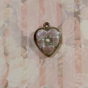May include: Heart-shaped pendant with a floral design. The pendant features a light pink flower with a pearl-like center, set within a silver-colored frame. The background has a floral pattern.