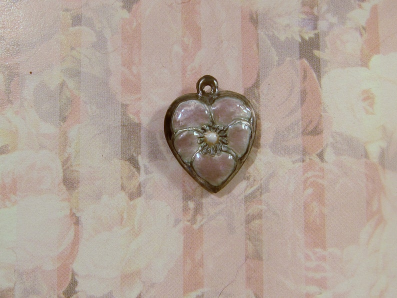 May include: Heart-shaped pendant with a floral design. The pendant features a light pink flower with a pearl-like center, set within a silver-colored frame. The background has a floral pattern.