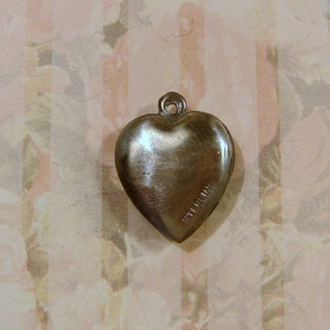 May include: A silver-colored heart-shaped pendant with a loop at the top for a chain. The word "STERLING" is stamped on the side of the heart. The pendant is likely made of sterling silver.