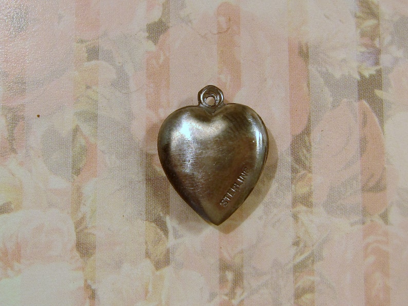 May include: A silver-colored heart-shaped pendant with a loop at the top for a chain. The word "STERLING" is stamped on the side of the heart. The pendant is likely made of sterling silver.