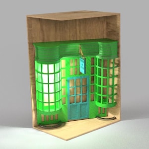 May include: A miniature green shop facade with a teal door and windows, set inside a wooden box. The shop has two cylindrical window displays with a grid pattern, and a small sign hangs above the door. A whimsical, decorative item.