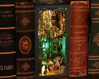 May include: A detailed, illuminated diorama of a room scene, set within a decorative black box, flanked by antique books. The interior features miniature figures, furniture, and a green-lit ambiance. The books have gold lettering and decorative covers.