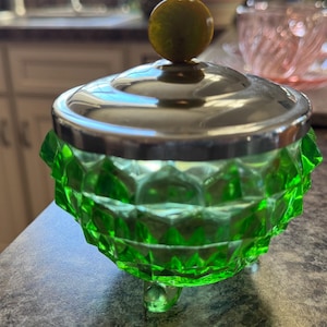 Könnte beinhalten: Eine grüne Glasschüssel mit einem silbernen Deckel und einem grünen Glasknopf. Die Schüssel hat ein Diamantenmuster und steht auf drei Beinen.