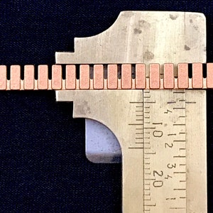 K&ouml;nnte beinhalten: Nahaufnahme eines Metalllineals, das eine kupferfarbene Kette misst. Das Lineal ist in Zoll und Millimetern gekennzeichnet. Die Kette besteht aus kleinen, rechteckigen Gliedern.