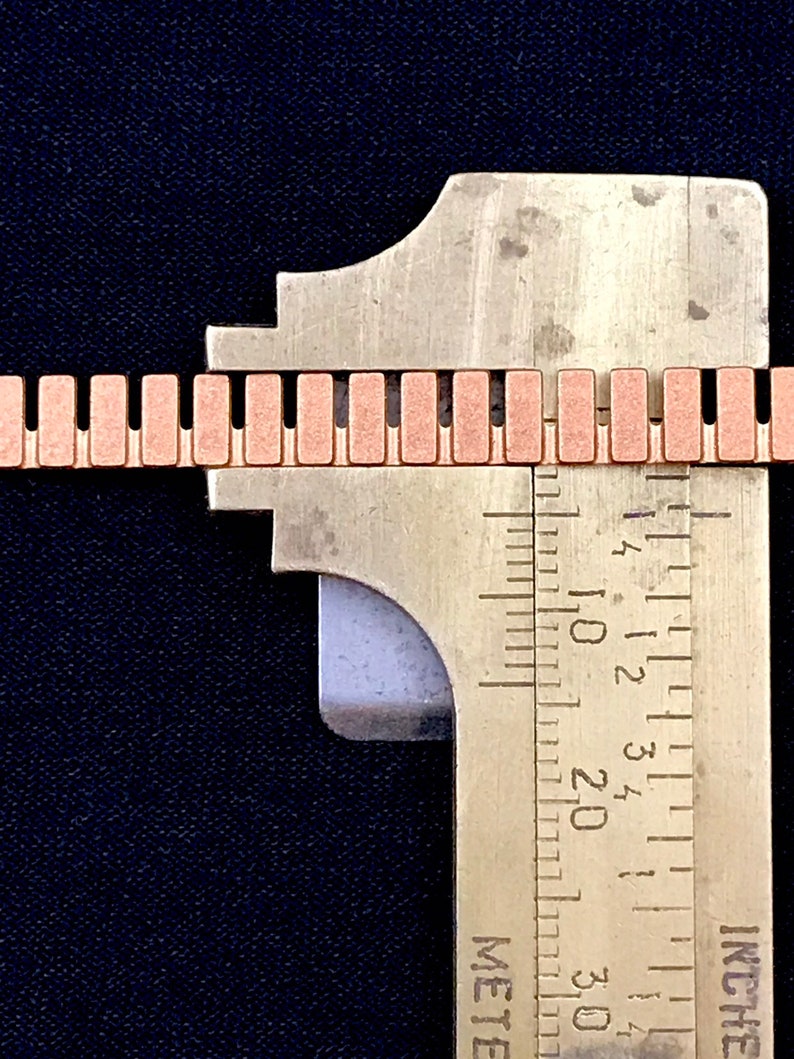 K&ouml;nnte beinhalten: Nahaufnahme eines Metalllineals, das eine kupferfarbene Kette misst. Das Lineal ist in Zoll und Millimetern gekennzeichnet. Die Kette besteht aus kleinen, rechteckigen Gliedern.