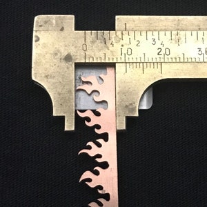 May include: A close-up of a metal ruler measuring a copper-colored metal strip with a flame design. The ruler shows measurements in inches.