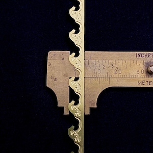 Puede incluir: Un ribete de metal dorado con un patrón de onda decorativo, medido con una regla en pulgadas.