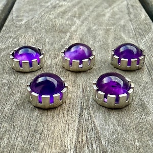 May include: Five amethyst cabochons set in silver-coloured metal bezels. The gemstones are a deep purple colour and have a rounded, polished surface. The bezels have a decorative, notched edge.