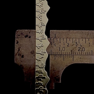May include: A gold metal strip with a repeating pattern of cactus silhouettes. The strip is measured with a ruler, showing it is approximately 5 cm long.