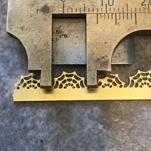 May include: A brass decorative piece with a spiderweb design is measured with a caliper. The caliper shows a measurement of approximately 0.4 inches. The piece has a repeating pattern of spiderwebs.