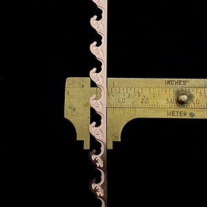 以下が含まれることがあります： ピンク色の装飾的なトリムのクローズアップで、縁が波打っています。トリムは定規で測定されており、約7.6cmの長さであることがわかります。