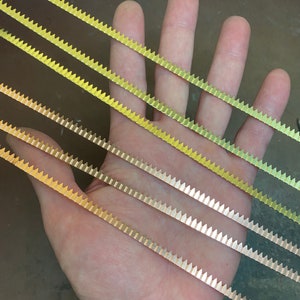 Może przedstawiać: Pięć pasków złotej metalowej listwy z ząbkowanym brzegiem. Listwa jest ułożona po przekątnej na dłoni.