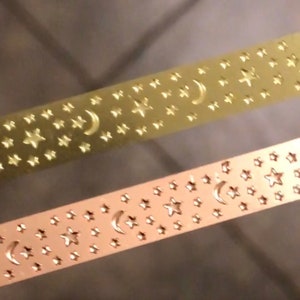 May include: Two rolls of gold-colored washi tape with a repeating pattern of stars and crescent moons.